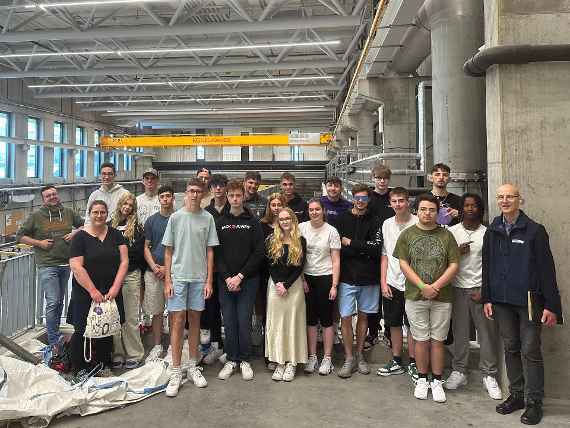 A group of students is standing in the main channel of the hydraulic engineering laboratory. In the background and foreground, one can see large model test channels. Eine Gruppe von Schüler:innen steht im Main Channel des Wasserbaulabors. Im Hintergrund und im Vordergrund sieht man große Modellversuchsrinnen.