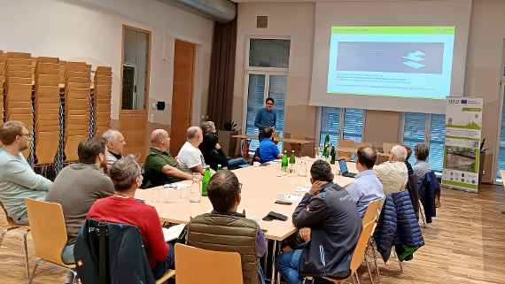 Die Teilnehmenden der Veranstaltung sitzen in einem Besprechungsraum. Eine Person hält mir Hilfe einer Präsentation einen Vortrag.