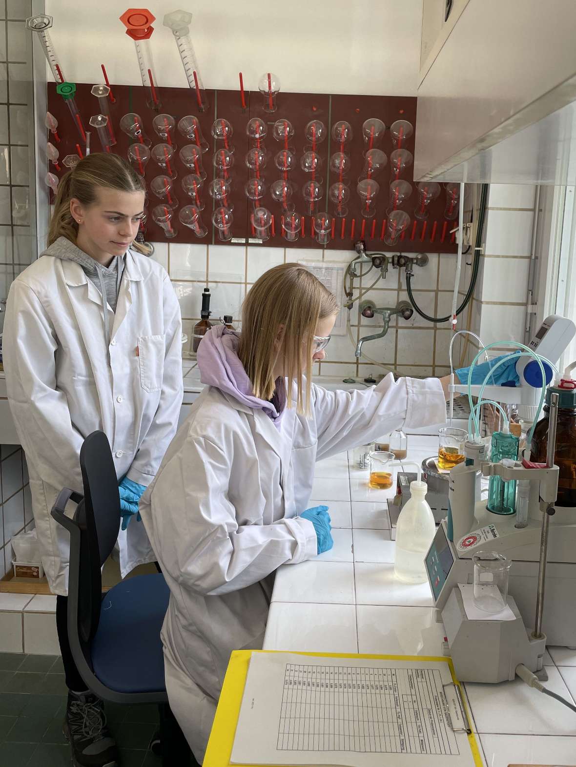 Zwei Teilnehmerinnen, bekleidet mit einem Laborkittel und blauen Handschuhen, befinden sich im Labor und bestimmen den Gelöst-Sauerstoff-Gehalt mittels Titration. Dazu werden verschiedene Gefäße verwendet.